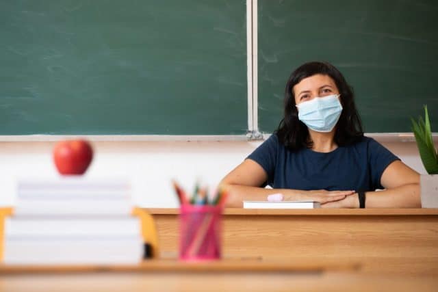 Decorative stock image shows a teacher at her desk wearing a mask.
