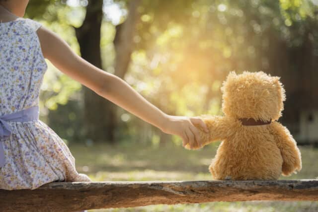 A child sits with her teddy bear. They are holding hands. 