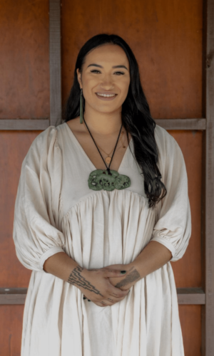 Jessica has long brown hair and is wearing a white flowing dress. She is wearing a pounamu taniwha necklace and earing.