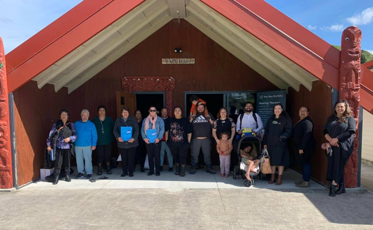 Several workshop participants, including adults and children, stand in front of a marae.